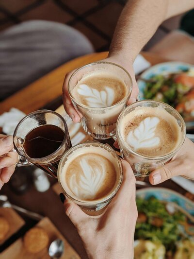 Four people clink glasses of latte and black coffee at a cozy coffee shop in Temple, Texas, with dishes in the background featuring intricate leafy latte art designs.