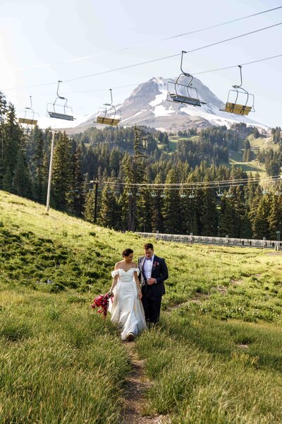 Groom is walking behind the bride. They are at a ski resort in the summer. They are among grass with trees behind. There is a ski lift above them and a mountain in the distance