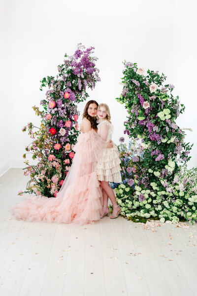 A woman in a flowing blush pink ruffled gown embraces a young girl in a cream lace dress, both smiling softly while standing between two lush floral arches filled with lilacs, roses, and greenery on a light wood floor scattered with petals.