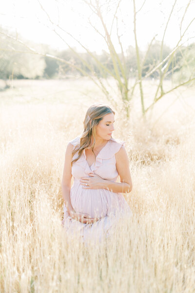 pregnant mom in pink dress holding belly sitting in tall grass in scottsdale arizona