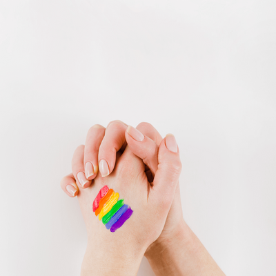 Two hands clasped together with rainbow pride colors painted on skin against white background, showing LGBTQIA+ support and solidarity