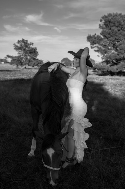 A lady in white dress and cowboy hat poses beside a grazing horse in a sunlit grassy field under a clear blue sky.