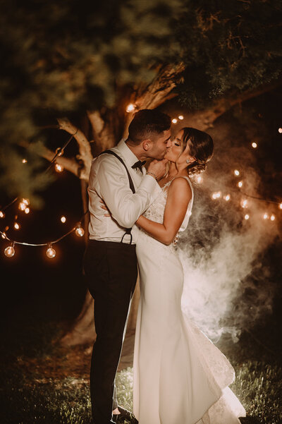 bride and groom kiss under tree with string lights at saltwater farm vineyard wedding photo by cait flethcer photography