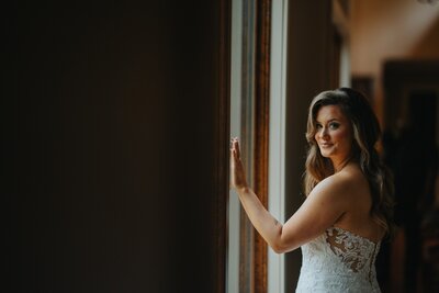 Bride standing in front of window with hands on glass, looking at the camera on her left, smirking.
