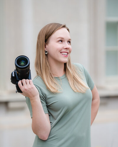 EmmiClaire photography's emily alwood holds her camera up smiling at something in the distance with her hand on her hip