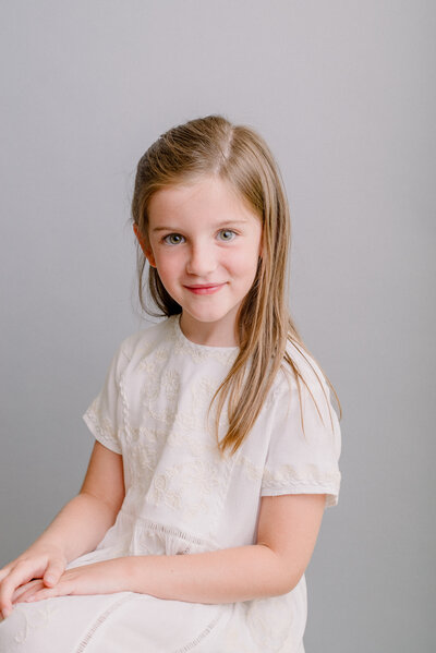 A first grade girl sits for her Redding CA school portraits in a white dress against a modern gray background.