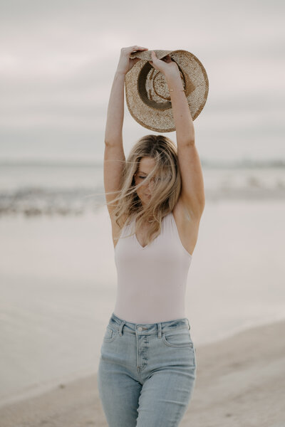 Woman holding a hat above her head on the beach for a portrait session with Tami Keehn