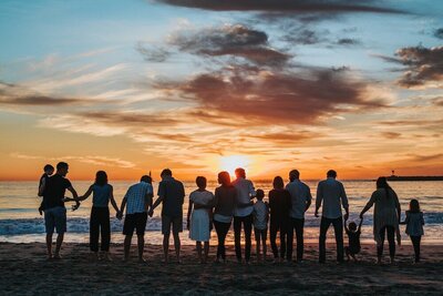 A large group of multi-generational people stand at the water's edge on a beach while holding hads and looking at the sunset with their backs to the camera.