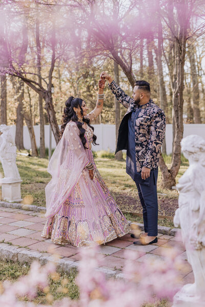 South Asian couple dancing beneath pink blossoms, the bride wearing a detailed pastel lehenga.