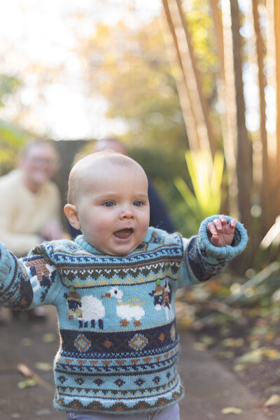 young toddler smiling as he runs at the camera parents can be seen out of focus behind him