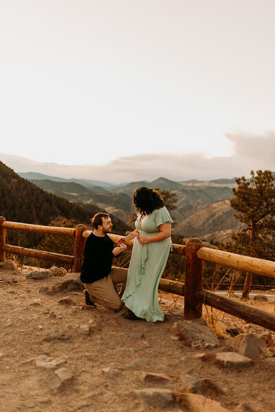 Groom on one knee proposes to girl in dress with wooden fence behind them overlooking Rocky Mountains