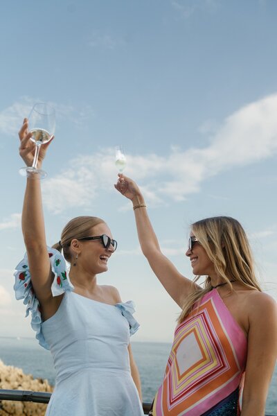 Two wedding guests holding up drinks with a blue sky and clouds in the background