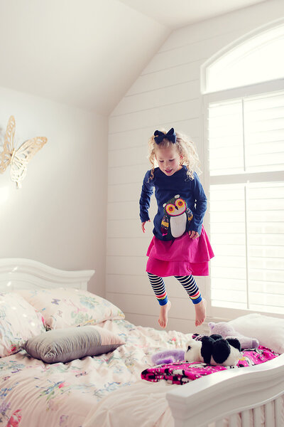Girls with curly hair in a blue bow and colorful outfit is jumping on her bed in Cincinnati, Ohio.