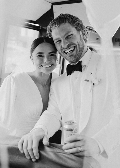 Black and white photo of bride wearing elegant long sleeve gown with hair slicked back next to groom in white tux and black bow tie while holding a beer