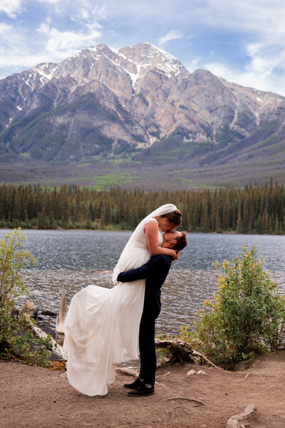Groom picks up Bride in front of mountain background in Pyramid Lake, Jasper, Alberta