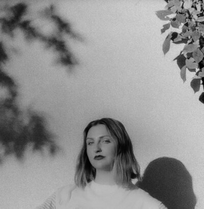 A black-and-white portrait of a woman standing outdoors, partially illuminated by natural light. The shadows of leaves frame the image, adding depth and texture to the background. The woman has shoulder-length hair and a confident expression, gazing directly at the camera.