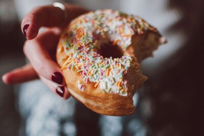 `A person holds a tempting doughnut with colorful sprinkles, partially bitten, against the blurred backdrop of a cozy coffee shop. Their fingernails are painted in rich dark red, adding to the charm of this Texas moment.W3