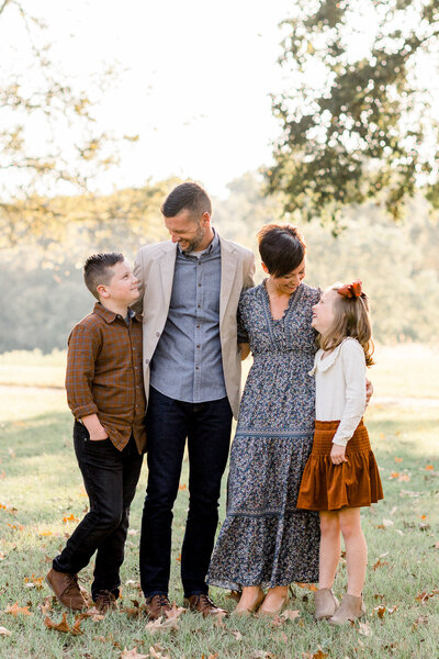 Outdoor fall family session with mom, dad, sister, and brother in muted neutral tones looking and smiling at each other with beautiful light shining through the trees behind them. Brandon, Mississippi Photographer