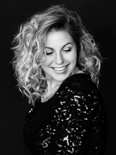 Black and white photo of a woman with curly hair smiling and gazing downward. They are wearing a sequined top and a necklace, with a dark background enhancing the elegant and joyful mood.