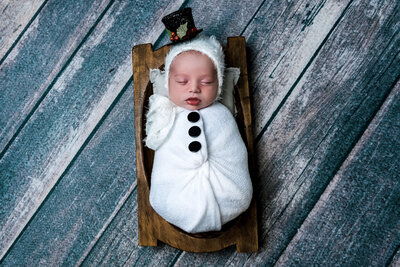 Connecticut newborn photographer creates whimsical winter portrait of sleeping baby dressed as snowman in white wrap with black buttons on wooden bed