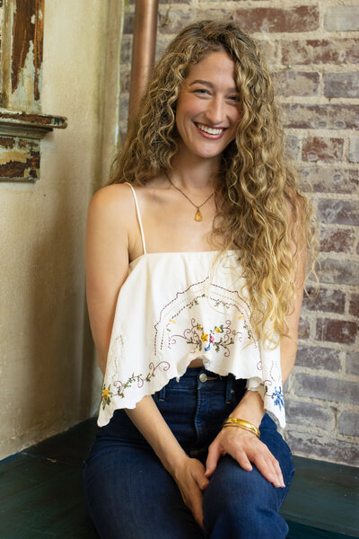 Dr. Mamie Burruss, ND, LAc smiling and sitting casually in front of exposed brick