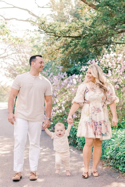 A family of 3 walks on a garden pathway holding hands during their family photos in NJ.