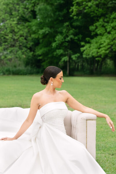 A wedding photo of a bride in a white dressing sitting sideways on a white sofa posing for a photo