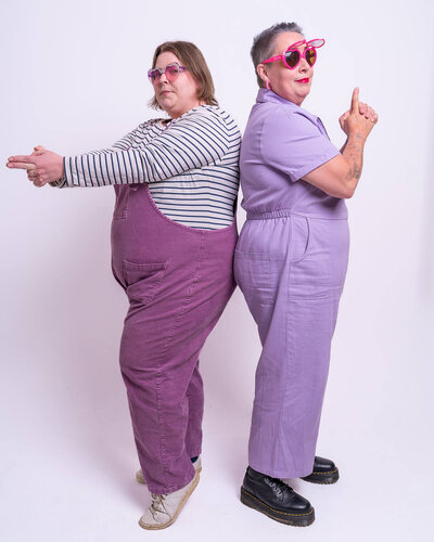 Two people, Lucy & Pippa, stand back-to-back in a studio, striking playful finger-gun poses – giving Austin Powers kinda vibes. Lucy left wears a striped top with mauve dungarees and pink sunglasses; Pippa right wears a lilac jumpsuit with bright pink heart-shaped sunglasses, black platform shoes, and visible forearm tattoos.