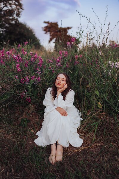 Young, red-headed woman sits in a flowy white dress in a field of flowers while closing her eyes and resting her head in her hands.