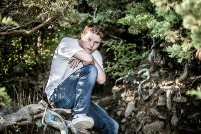 A high school senior boy wearing a white t-shirt & jeans, sitting on a tree trunk with his arms crossed posing for a south shore ma senior photographer