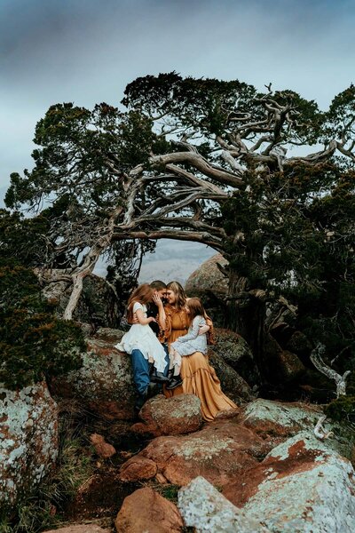 In front of a misshaped tree, a family of four cuddles close together, with the mother and father face-to-face, and the daughters in the parent's laps.