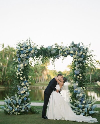 Bride and groom standing beneath a white and blue hydrangea floral arbor at their luxury coastal wedding at Fairchild Tropical Botanic Garden in Miami.