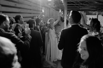Bride and wedding party raising champagne glasses in a celebratory toast, captured in candid black and white.