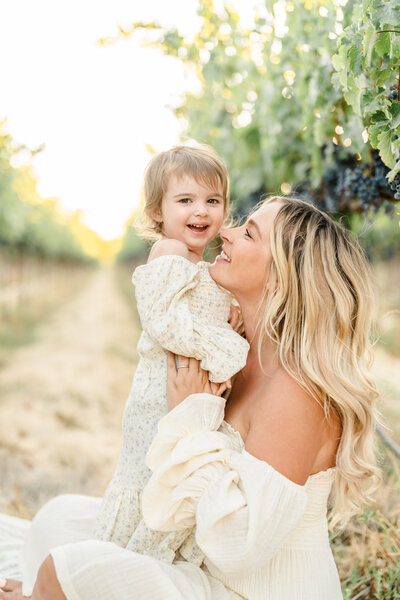 Mother sits and holds her daughter as she stands on her lap in an orchard, photography from Bay area family photographer Light Livin Photography
