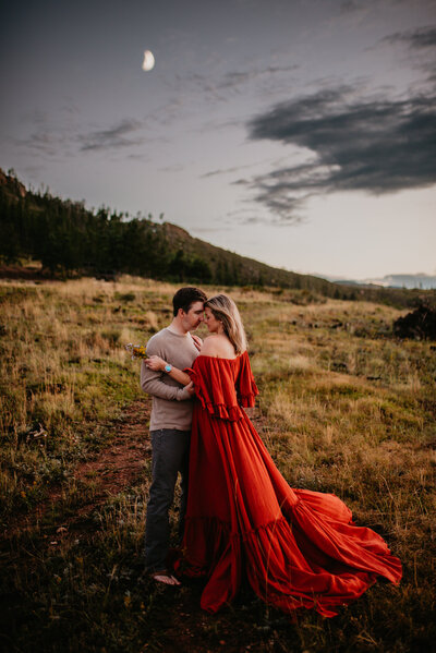 An engaged couple embraces under the moon as the sun starts to set on top of a mountain in Rist Canyon