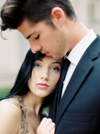 Blue eyed and black haired bride with tattoos and berry lips looks into the camera while leaning on the chest of her groom wearing a black suit in Spain.