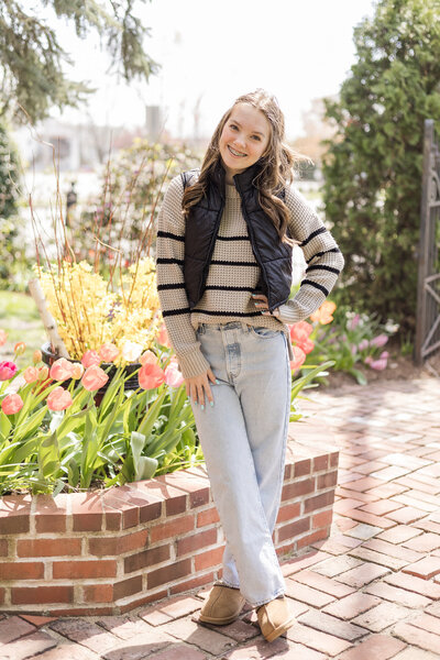 high school senior female, standing on a brick path with flowers behind her, wearing blue jeans, a light tan long sleeve shirt a black zip up jacket and smiling at the camera
