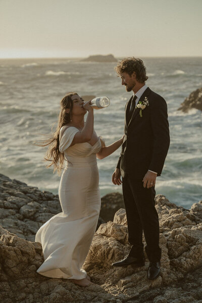 big sur elopement photo of a bride with a bottle of champagne and her groom looking at her while standing on the coast of garrapata state park in big sur california