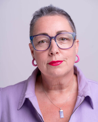 Pippa from Disobedient Business® Co. looks confidently at the camera, wearing purple glasses, pink hoop earrings, and a lilac collared boiler suit, with bright pink lipstick and a calm, self-assured expression against a light background.
