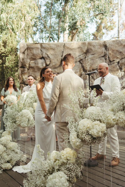 Young couple getting married at Calamigos Ranch at the Oak Room by friend in white suit with hydrangeas and babies breath surrounding couple 