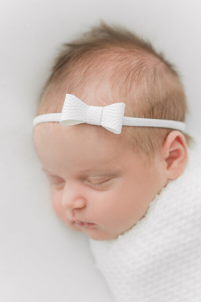 A closeup photo of a newborn baby girl sleeping with a white bow headband on her head at a DC Newborn Photography session