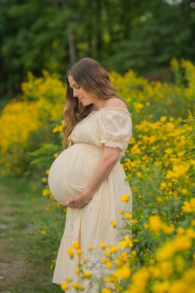 pregnant mom in white crop top and jeams holding a white and yellow boquet leaning on the wall with her husband as he gazes at her