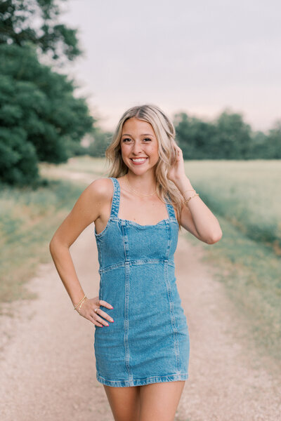 Senior picture of girl in denim dress walking on park trail by field