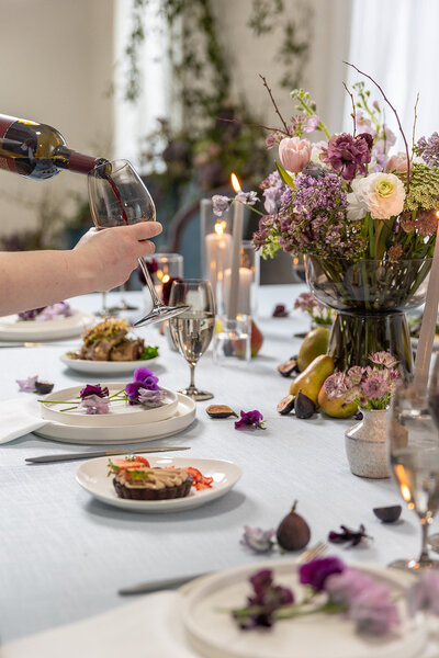 Wedding table set up with a white table clothe, purple florals, and gourmet food. Someone is pouring a glass of red wine