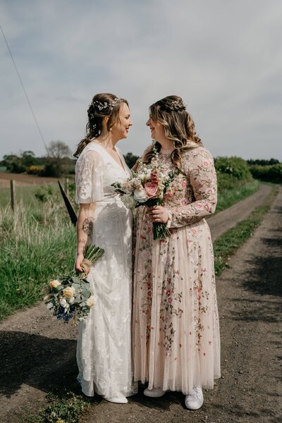 An LGBTQ+ couple candidly pose together for wedding photography as they walk down a country path. They walk hand in hand.