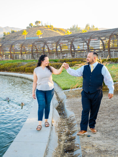 A couple smiles at each other and he holds up her hand gently as she walks along the bordering concrete of a duckpond with a artistic bridge behind them. Photo by SAVI Photography - Photographer in Riverside