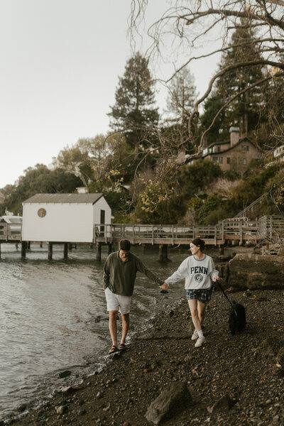 Couple holding hands and walking dog down rocky coastal beach with dock in the background