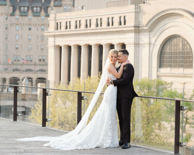 bride with long veil embracing groom in dark suit outside NAC wedding venue.  Captured by Ottawa wedding photographer JEMMAN Photography