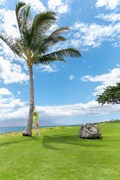 Wide shot of palm tree with girl leaning on it, with open sky and lush green grass.