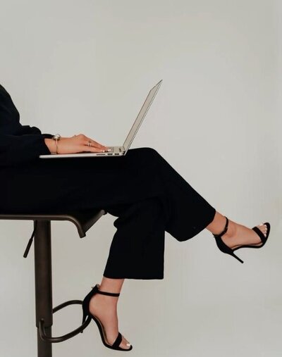 A minimalist workspace featuring a light wooden desk and a black leather chair. On the desk are a cream lamp, a beige candle, and an open MacBook. Natural daylight casts soft shadows on the wall.